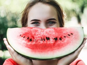 Phot of a Caucasian female teen smiling which she holds up a watermelon slice in front of her face that is supposed to look like her smile. Photo could represent the happiness and confidence she now feels since working with a solution focused brief therapist in Illinois proving online anxiety treatment for teens.