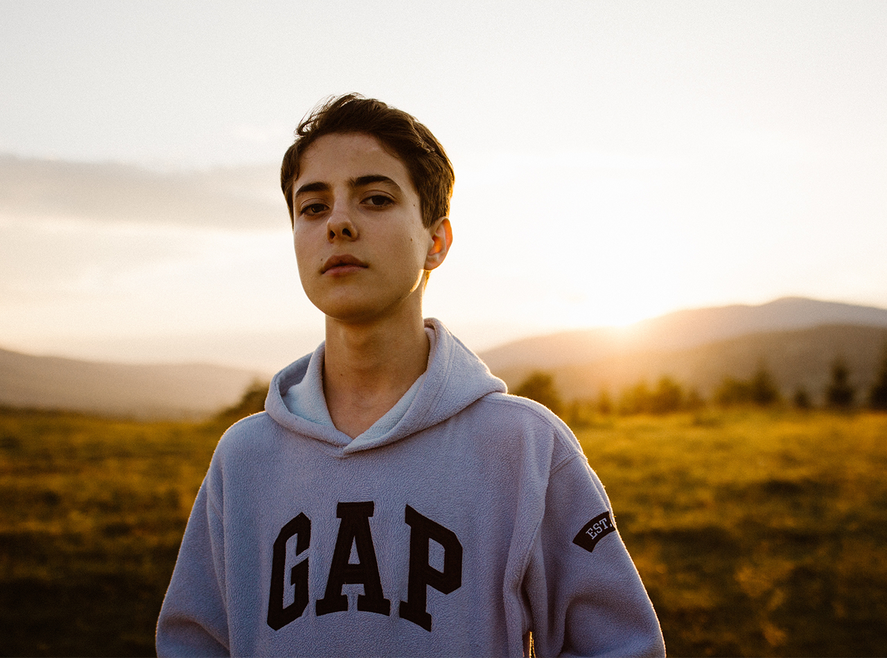 Photo of an unsmiling Caucasian male teen wearing a GAP hoodie and standing outside with the sun setting behind him. Photo could represent a stressed and anxious teen and how receiving online therapy for anxiety in Illinois might be helpful.