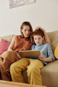 Photo of Caucasian mother sitting on a couch with her redheaded son with her hand touching his head and both looking at the ipad on the son's lap which could represent them having an online counseling session in Illinois for her son's anxiety due to loss.