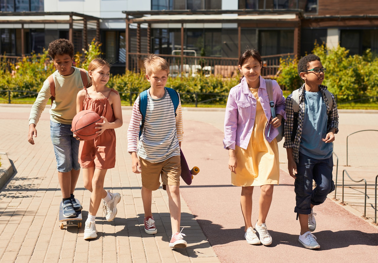 Group of middle school kids of different races walking out of school together on a summer afternoon with their school building behind them. Photo could represent how they're all getting along and becoming friends regardless of race or socioeconomic status.