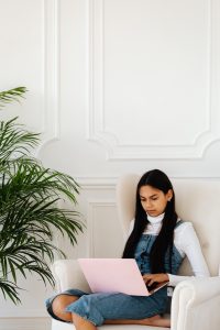 Photo of a young Latina wearing a white turtleneck sweater with a jeans jumper sitting in a white overstuffed chair with her pink iPad on her lap and a floor plant to her left. Photo could represent an anxious teens having online anxiety treatment with her online solution focused brief therapist in Illinois