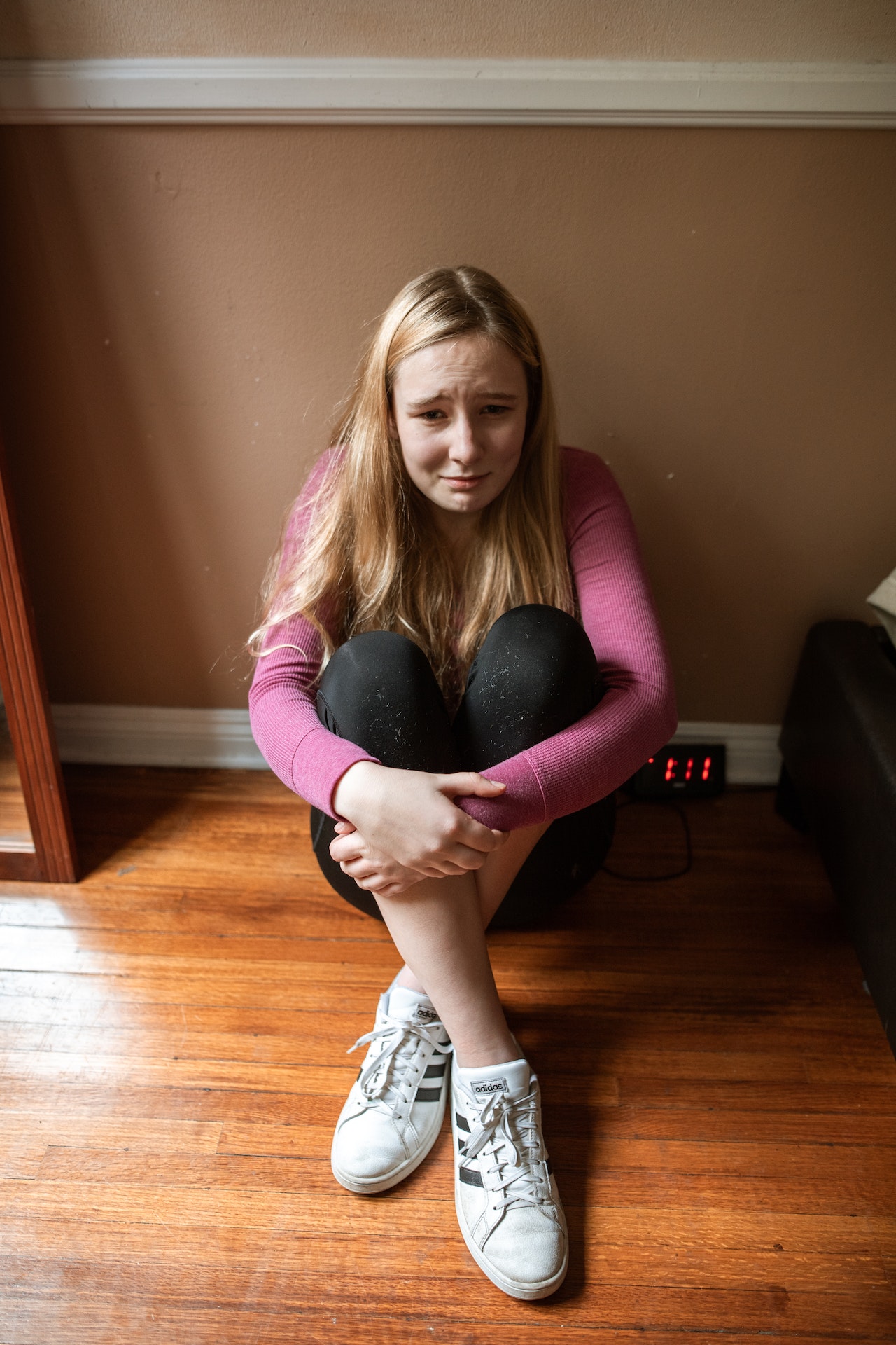 Photo of Caucasian teen female blond sitting on the floor with her knees up in front of her and her hands clasped together on them. She has a worried and tearful look on her face. Photo could represent the stress she feels from having anxiety and the need to see an online therapist in Illinois for anxiety treatment.
