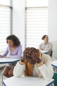 Photo of African American teen male sitting in a desk in a classroom with his hands on his head while staring down at the paper on his desk. Photo could represent test anxiety and the need for online solution focused brief therapy in Illinois.