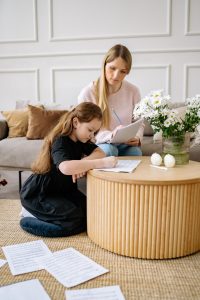 Photo of Caucasian girl with long brown hair sitting on her knees in front of a wood table writing on a piece of paper while her mother sits beside her on a couch also holding papers and looking over her daughter's shoulder. Photo could represent the test anxiety daughter is feeling so they are doing practice exams together to ease her fears. Daughter would likely benefit from anxiety treatment for kids from an online solution focused brief therapist in Illinois.