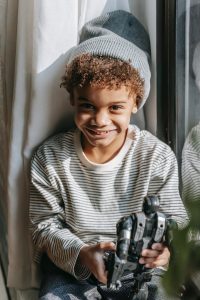 Photo of smiling African American boy wearing a knitted hat and striped shirt while holding a toy in his hands. Photo could represent how much better he feels after working through his anxiety and behavioral issues with his online therapist for kids in Illinois.