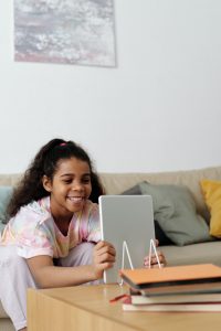 Photo of African American girl seated on a beige couch smiling and holding her iPad in her hands while leaning forward on the coffee table. Photo could represent an online therapy session with her online solution focused brief therapist in Illinois for anxiety treatment.
