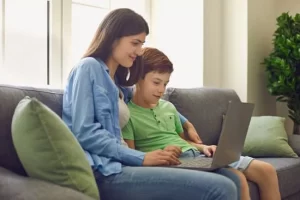 Photo of Caucasian Mom and her redheaded son sitting on a blue couch together with the mom's arm wrapped around the son. They are both smiling as they look at the open laptop in front of them on their laps. Photo could represent an online session with a Christian therapist in Illinois to help the son with his anxiety.