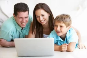 Photo of Caucasian family smiling as they all gather around the laptop looking into the screen. Photo could represent a therapy session with the boy's online solution focused brief therapist in Illinois for anxiety treatment.