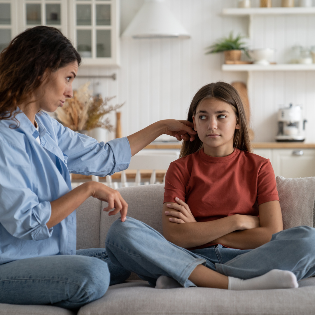 Photo of Caucasian girl and mother sitting on couch together. The daughter's arms are crossed and she is giving a side eye to her mom who looks concerned. Photo could represent teen anxiety and the need for online solution focused brief counseling for teens in Illinois and Florida.