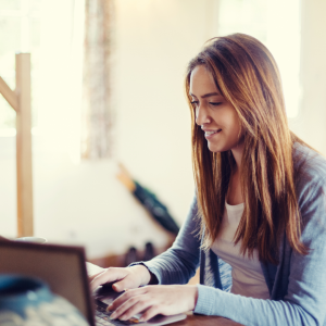Photo of Caucasian female teen sitting at desk with her hands on the keyboard and smiling in to the screen. Photo could represent an online Christian counseling session for teens in Illinois.