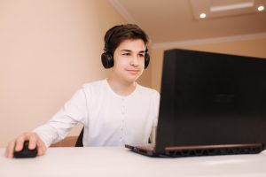 Photo of Caucasian male teen sitting at his desk with headphones on and smiling into his laptop screen. Photo could represent him having online solution focused brief therapy with his online Christian counselor in Illinois who specializes in kids and teens with anxiety.