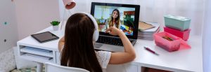 Photo of a girl from behind sitting at a white desk with headphones and engaging with a smiling woman on the screen in front of her on the lap top. Photo could represent an online therapy session with her Christian therapist in Illinois who specialized in kids and teens with anxiety.