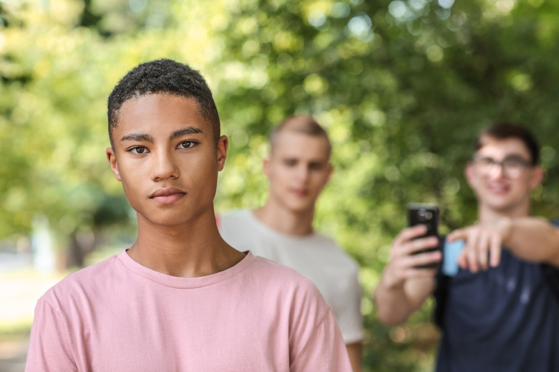 Photo of African American teen standing outside and looking into the camera while two male teens are behind him, pointing their phones at him and laughing. Photo could represent the peer pressure and anxiety he feels resulting in the need for Christian counseling for anxiety in teens in Illinois and Florida.