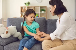 Photo of Caucasian mother and young daughter sitting on a grey couch as they face each other holding hands and the daughter looking down a bit sad. Photo could represent the benefit of open communication between the parent and child in helping her with her social anxiety. Photo could also represent the need for additional support in the way of online Christian counseling for kids and teens with anxiety in Illinois and Florida.